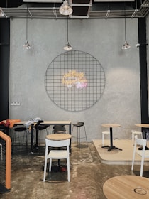 A modern cafe interior with industrial design elements featuring exposed ceilings and concrete walls. A circular metal grid dominates the wall with a neon sign at its center. Several chairs and tables are scattered around the room, with a variety of seating options including stools and regular chairs. Hanging pendant lights provide illumination.