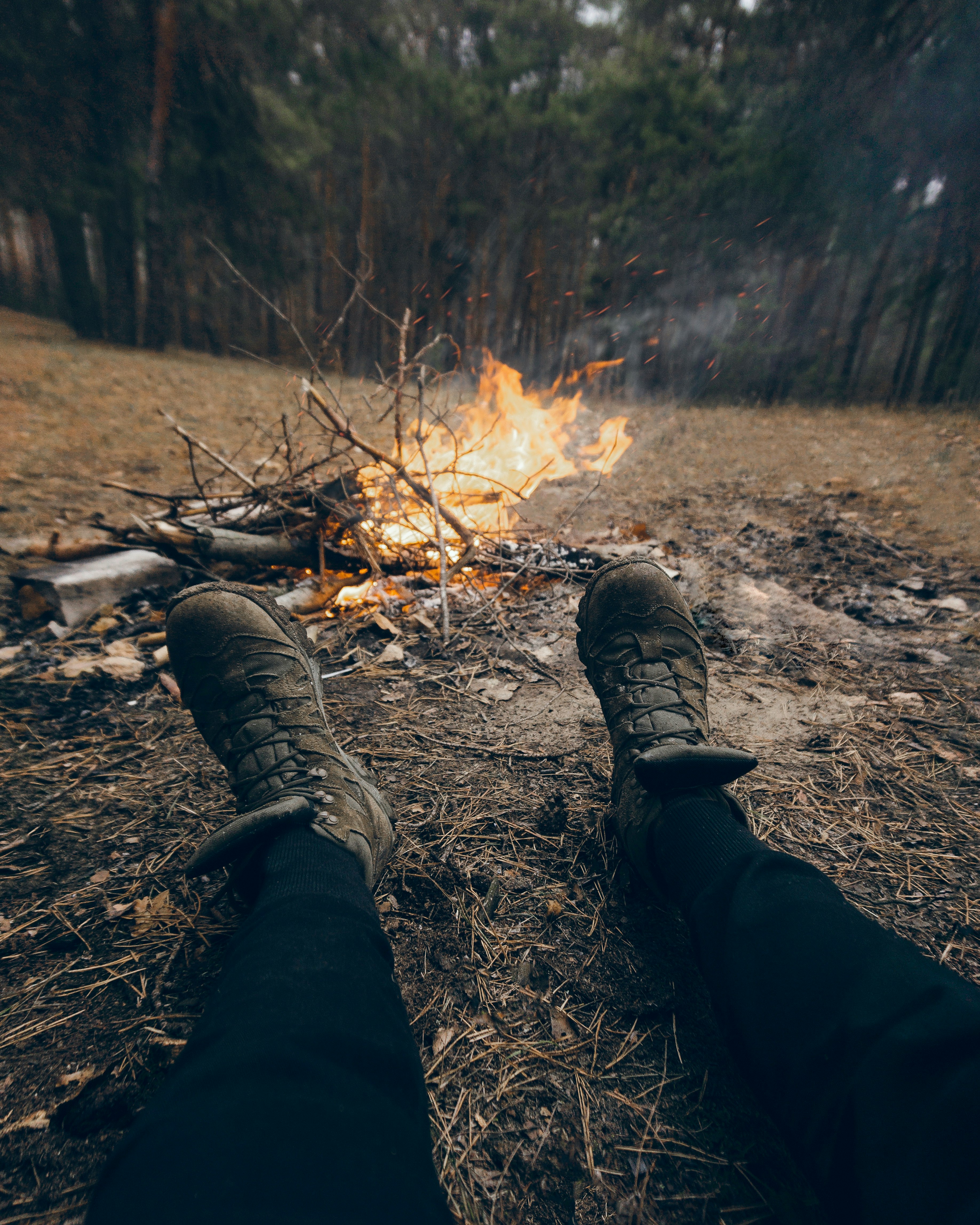 person in black pants and brown hiking boots sitting on ground near bonfire during daytime