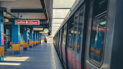A metro station platform with a train on the right side and a sign indicating a ladies-only section in both English and Arabic. The platform is lined with blue and yellow pillars, and a clock hangs from the ceiling.