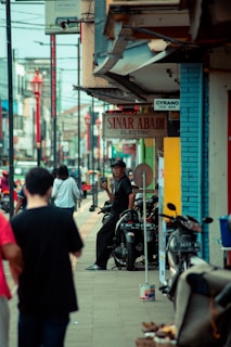 A busy urban street scene with several people walking along the sidewalk. A man is sitting on a motorcycle, holding a phone while smiling. Various shops and signs, such as 'Sinar Abadi Electric', are visible. The atmosphere is lively, with a mix of pedestrians and motorcycles lining the street.