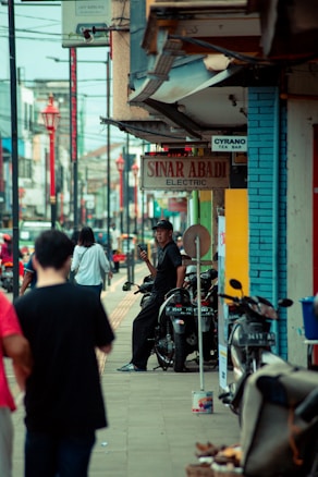 A busy urban street scene with several people walking along the sidewalk. A man is sitting on a motorcycle, holding a phone while smiling. Various shops and signs, such as 'Sinar Abadi Electric', are visible. The atmosphere is lively, with a mix of pedestrians and motorcycles lining the street.