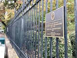 An ornate black iron fence runs the length of a stone wall, with trees and foliage visible behind it. A bronze plaque is mounted on the fence, providing historical information about the Georgetown Reservoir site. The leaves on the trees display autumn colors, with hints of green, yellow, and orange.