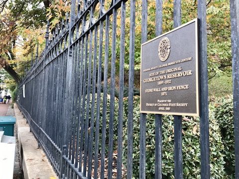 An ornate black iron fence runs the length of a stone wall, with trees and foliage visible behind it. A bronze plaque is mounted on the fence, providing historical information about the Georgetown Reservoir site. The leaves on the trees display autumn colors, with hints of green, yellow, and orange.