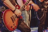 A person is playing an electric guitar with a sunburst finish. The guitar is being held firmly with the left hand on the neck, wearing a bracelet and a ring. Several people stand in the background, slightly out of focus, suggesting a lively atmosphere, possibly on stage.