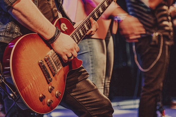 A person is playing an electric guitar with a sunburst finish. The guitar is being held firmly with the left hand on the neck, wearing a bracelet and a ring. Several people stand in the background, slightly out of focus, suggesting a lively atmosphere, possibly on stage.