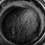 A close-up view of a jar filled with a fine, granular, grayish-black powder. The texture appears coarse with small, uneven particles piled within the container. The image presents minimal lighting, enhancing the dark shade and texture of the powder.