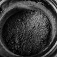 A close-up view of a jar filled with a fine, granular, grayish-black powder. The texture appears coarse with small, uneven particles piled within the container. The image presents minimal lighting, enhancing the dark shade and texture of the powder.