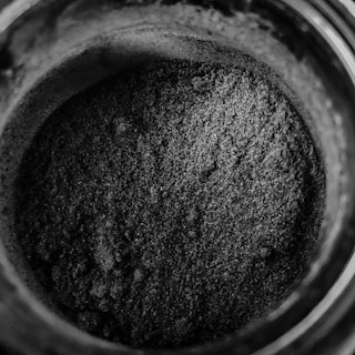 A close-up view of a jar filled with a fine, granular, grayish-black powder. The texture appears coarse with small, uneven particles piled within the container. The image presents minimal lighting, enhancing the dark shade and texture of the powder.