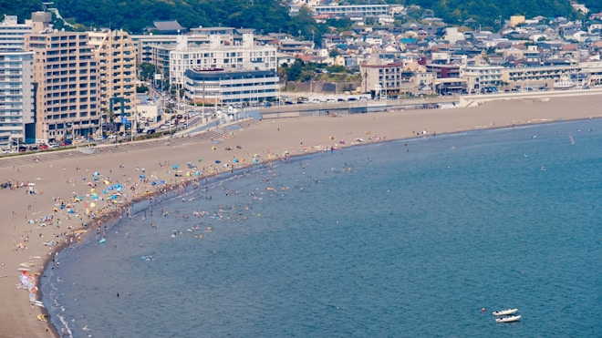 Aerial view of a crowded beach with numerous people enjoying the water and sand. Colorful umbrellas are scattered across the beach. In the background, a row of mid-rise buildings lines the waterfront.
