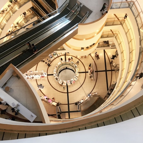 A modern shopping mall with multiple floors and an open central atrium. The view from above reveals circular architectural elements and escalators connecting the levels. There are several small kiosks on the ground floor surrounded by people.