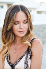 A young woman with long wavy brown hair and subtle makeup is looking downward. She wears a delicate camisole with black lace detailing and a simple necklace. The background is softly blurred, suggesting an outdoor setting.