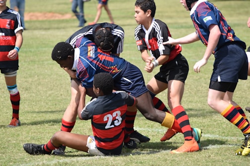 A group of young rugby players are in the midst of a game on a grassy field. They are wearing colorful uniforms, with one team in black and red and the other in blue and red. The players are engaged in a tackle, displaying teamwork and athleticism.