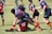 A group of young rugby players are in the midst of a game on a grassy field. They are wearing colorful uniforms, with one team in black and red and the other in blue and red. The players are engaged in a tackle, displaying teamwork and athleticism.