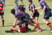 A group of young rugby players are in the midst of a game on a grassy field. They are wearing colorful uniforms, with one team in black and red and the other in blue and red. The players are engaged in a tackle, displaying teamwork and athleticism.