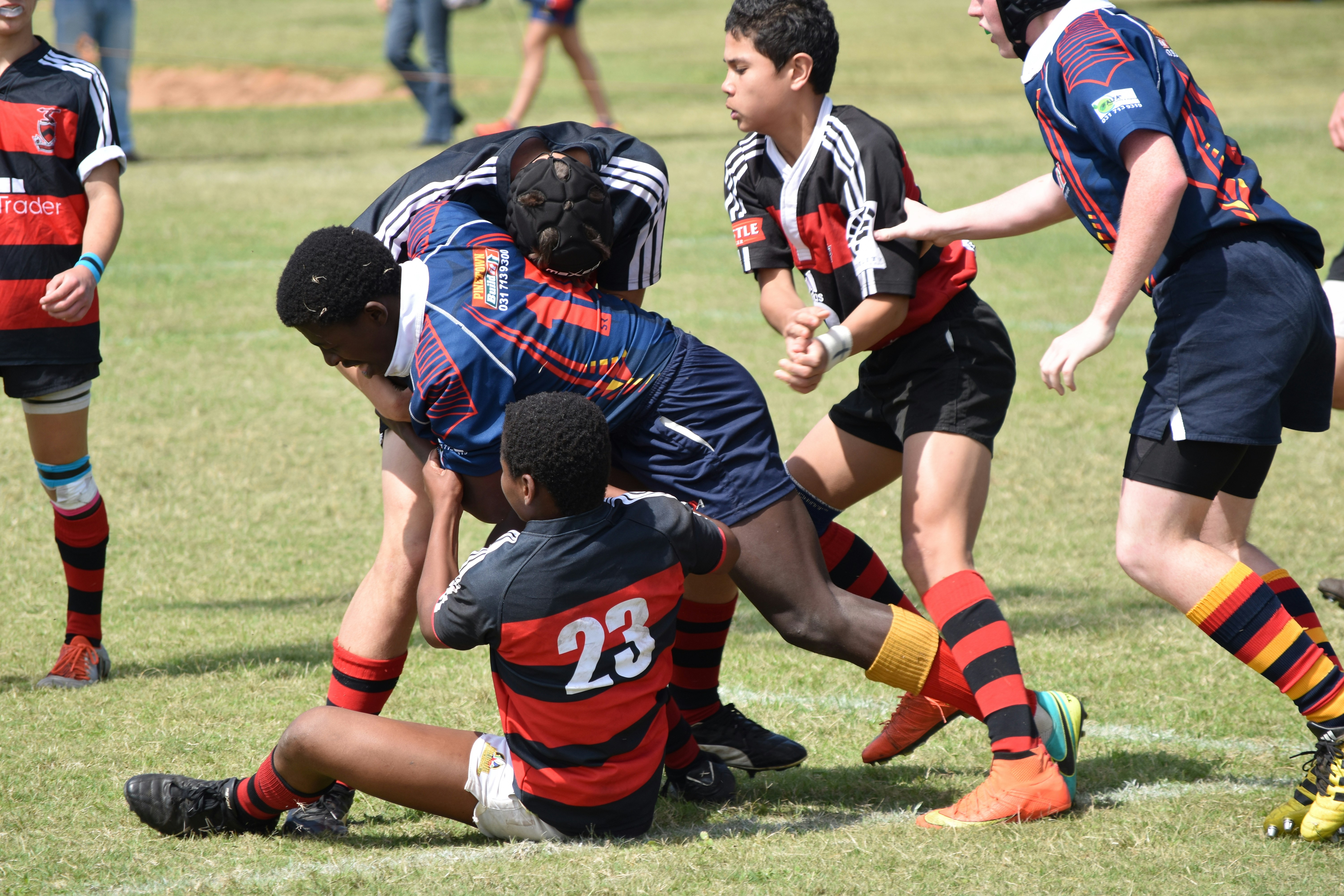 A determined youth, clutching a rugby ball pushes his way through his adversary's to gain ground, despite the other team tackling him and pulling him back.