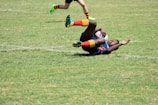 A rugby player in action, lying on the grass, holding a rugby ball tightly while another player's legs are visible in the frame running. The player on the ground is wearing brightly colored socks and a jersey.