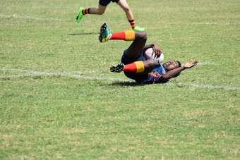 A rugby player in action, lying on the grass, holding a rugby ball tightly while another player's legs are visible in the frame running. The player on the ground is wearing brightly colored socks and a jersey.