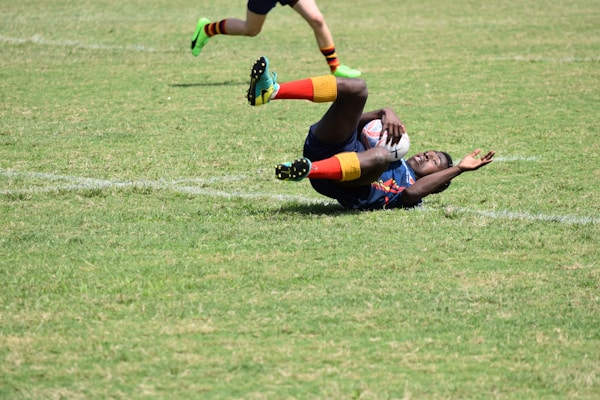 A rugby player in action, lying on the grass, holding a rugby ball tightly while another player's legs are visible in the frame running. The player on the ground is wearing brightly colored socks and a jersey.