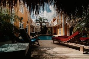 A tropical courtyard with a pool is surrounded by yellow buildings with white trim. Lounge chairs with red and blue cushions are positioned on a wooden deck, shaded by overhanging palm leaves. The sky is partly cloudy, providing a mixture of sunlight and shade.