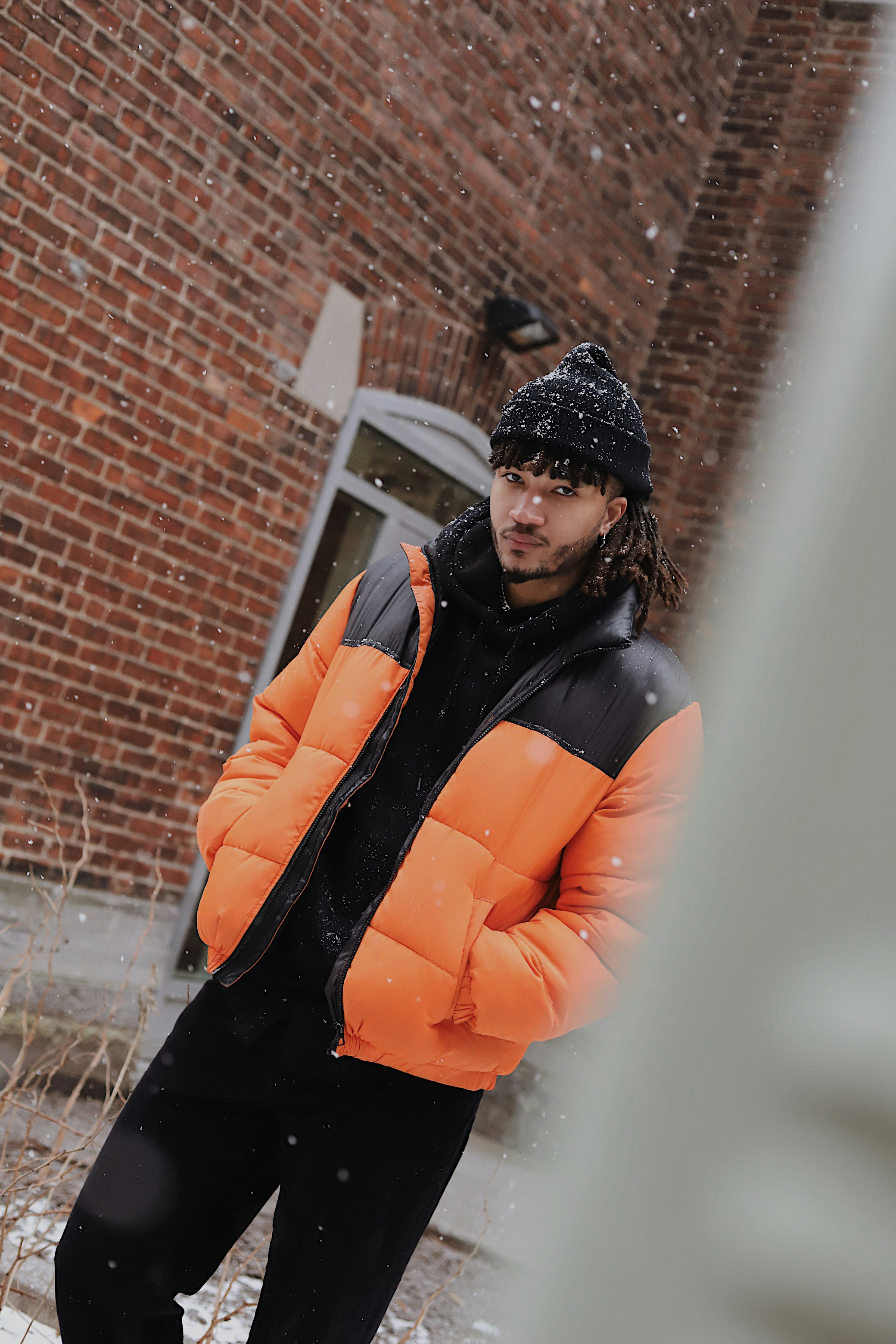 woman in orange jacket and black knit cap standing near brown brick wall during daytime