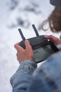 A person is holding a black remote control device with two antennas, likely used for operating a drone. The person is wearing a denim jacket, and the background appears snowy, conveying a cold environment.