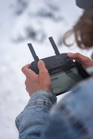 A person is holding a black remote control device with two antennas, likely used for operating a drone. The person is wearing a denim jacket, and the background appears snowy, conveying a cold environment.