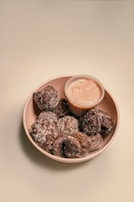 A bowl filled with small, sugar-coated chocolate pastries is presented alongside a small container of light-colored dipping sauce. The pastries have a textured surface and are arranged closely together.