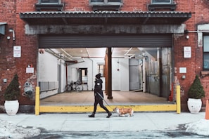 A person walks a dog past an open garage in an urban setting. The building features red brick walls and a large open shutter revealing an empty space inside with a bike. Yellow posts and potted plants are positioned next to the entrance, and no parking signs are visible.