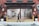 A person walks a dog past an open garage in an urban setting. The building features red brick walls and a large open shutter revealing an empty space inside with a bike. Yellow posts and potted plants are positioned next to the entrance, and no parking signs are visible.