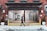 A person walks a dog past an open garage in an urban setting. The building features red brick walls and a large open shutter revealing an empty space inside with a bike. Yellow posts and potted plants are positioned next to the entrance, and no parking signs are visible.