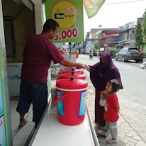 A man at a juice stand interacts with a woman and a small child on the street. The stand features large red containers labeled 'Jus' and a price sign overhead. The woman is handing something to the man, likely making a purchase.