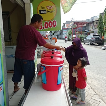 A man at a juice stand interacts with a woman and a small child on the street. The stand features large red containers labeled 'Jus' and a price sign overhead. The woman is handing something to the man, likely making a purchase.