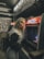 A person with long blonde hair wearing a black and white jacket stands beside an old arcade game machine in a dimly lit retro-style arcade. The game machine has a colorful retro design, and the setting has a nostalgic, vintage feel.