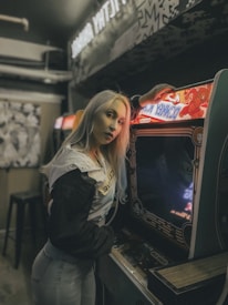 A person with long blonde hair wearing a black and white jacket stands beside an old arcade game machine in a dimly lit retro-style arcade. The game machine has a colorful retro design, and the setting has a nostalgic, vintage feel.