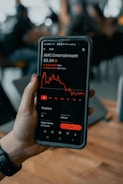 A hand holding a smartphone displaying a stock trading app screen. The app shows stock information for AMC Entertainment, including a line graph of its performance, current price, and trading options.