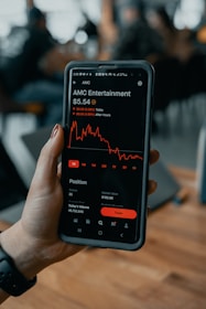 A hand holding a smartphone displaying a stock trading app screen. The app shows stock information for AMC Entertainment, including a line graph of its performance, current price, and trading options.
