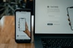 A smartphone and a laptop are displayed on a wooden surface. The smartphone screen shows an app interface featuring a financial portfolio, with stock symbols and numerical values. The laptop screen shows a website login page with promotional text offering incentives for signing up.
