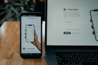 A smartphone and a laptop are displayed on a wooden surface. The smartphone screen shows an app interface featuring a financial portfolio, with stock symbols and numerical values. The laptop screen shows a website login page with promotional text offering incentives for signing up.