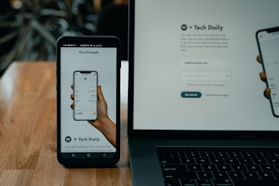 A smartphone and a laptop are displayed on a wooden surface. The smartphone screen shows an app interface featuring a financial portfolio, with stock symbols and numerical values. The laptop screen shows a website login page with promotional text offering incentives for signing up.