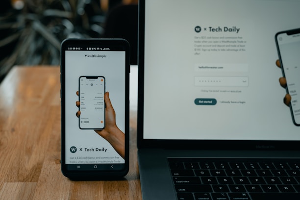 A smartphone and a laptop are displayed on a wooden surface. The smartphone screen shows an app interface featuring a financial portfolio, with stock symbols and numerical values. The laptop screen shows a website login page with promotional text offering incentives for signing up.