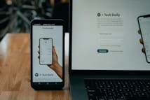 A smartphone and a laptop are displayed on a wooden surface. The smartphone screen shows an app interface featuring a financial portfolio, with stock symbols and numerical values. The laptop screen shows a website login page with promotional text offering incentives for signing up.