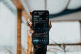 A hand is holding a smartphone displaying a stock market app. The screen shows financial data, including stock prices, a line graph, and trading volume. The background is blurred, featuring some indistinct indoor elements with natural light coming through.