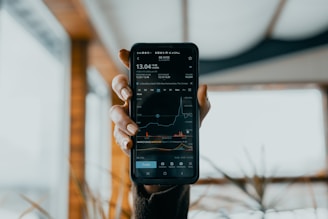 A hand is holding a smartphone displaying a stock market app. The screen shows financial data, including stock prices, a line graph, and trading volume. The background is blurred, featuring some indistinct indoor elements with natural light coming through.