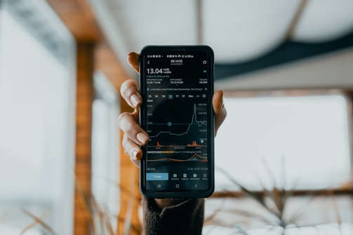 A hand is holding a smartphone displaying a stock market app. The screen shows financial data, including stock prices, a line graph, and trading volume. The background is blurred, featuring some indistinct indoor elements with natural light coming through.