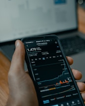 A person holds a smartphone displaying a stock market application with graphs and financial data. A laptop is visible in the background, slightly blurred.