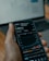 A person holds a smartphone displaying a stock market application with graphs and financial data. A laptop is visible in the background, slightly blurred.