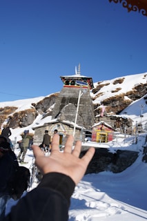 A temple stands prominently in a snowy mountainous landscape with clear blue skies above. Several people are walking up to the entrance, and a hand is extended towards the temple in the foreground. The temple is built with stone and has colorful accent decorations.