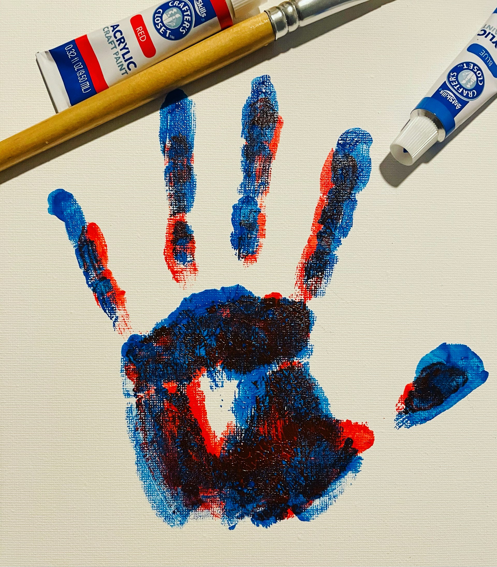 A colorful handprint created with red, blue, and hints of black paint is displayed on a white canvas. Beside the handprint are a wooden-handled paintbrush and tubes of red and blue acrylic paint.
