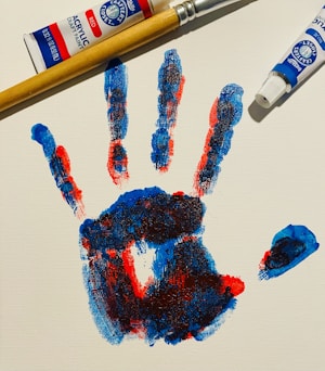 A colorful handprint created with red, blue, and hints of black paint is displayed on a white canvas. Beside the handprint are a wooden-handled paintbrush and tubes of red and blue acrylic paint.