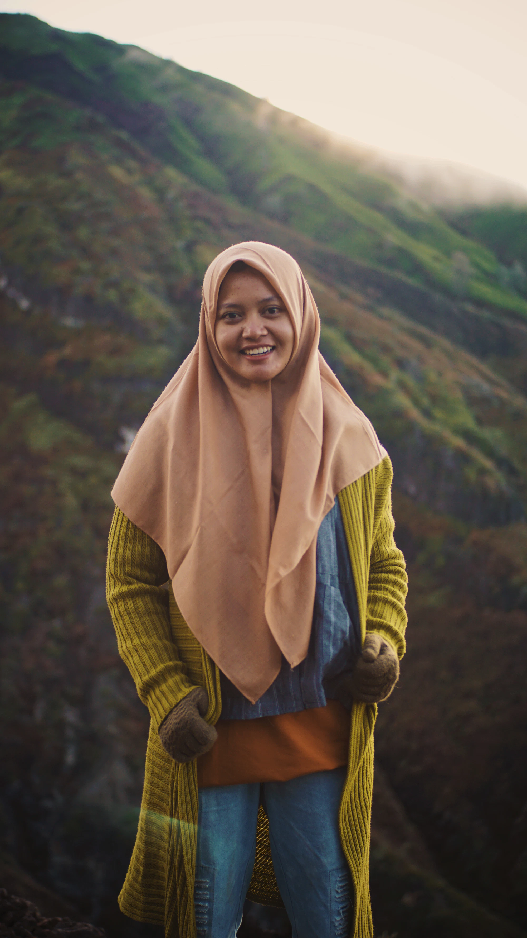 Perempuan berhijab tersenyum di lanskap perbukitan yang hijau.